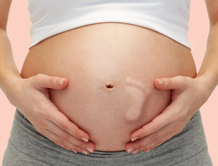 pregnancy, motherhood, people and expectation concept - close up of pregnant woman touching her bare tummy with kicking baby footprint over pink background